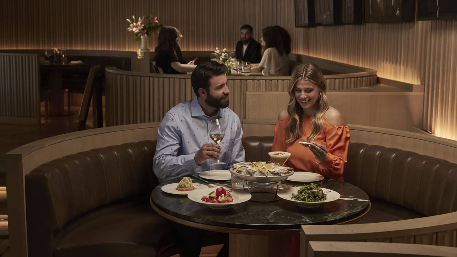 A couple sits at a round table in a leather banquette smiling and eating a plate of oysters and various other side dishes