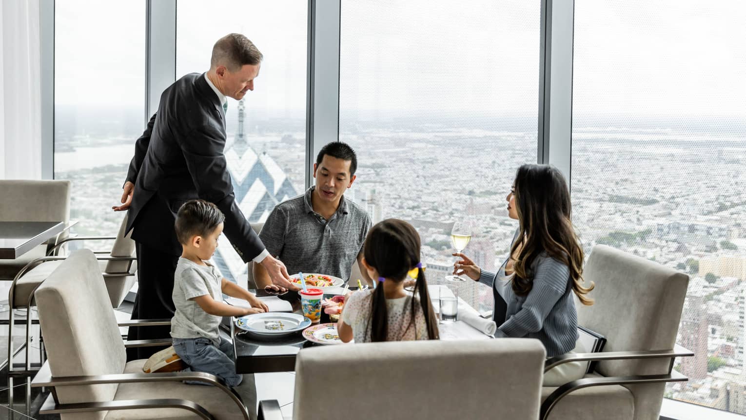 A staff member in a suit placing food on a table for a family of four including a mother, father, daughter, and son; there are large windows looking at a city below around them.