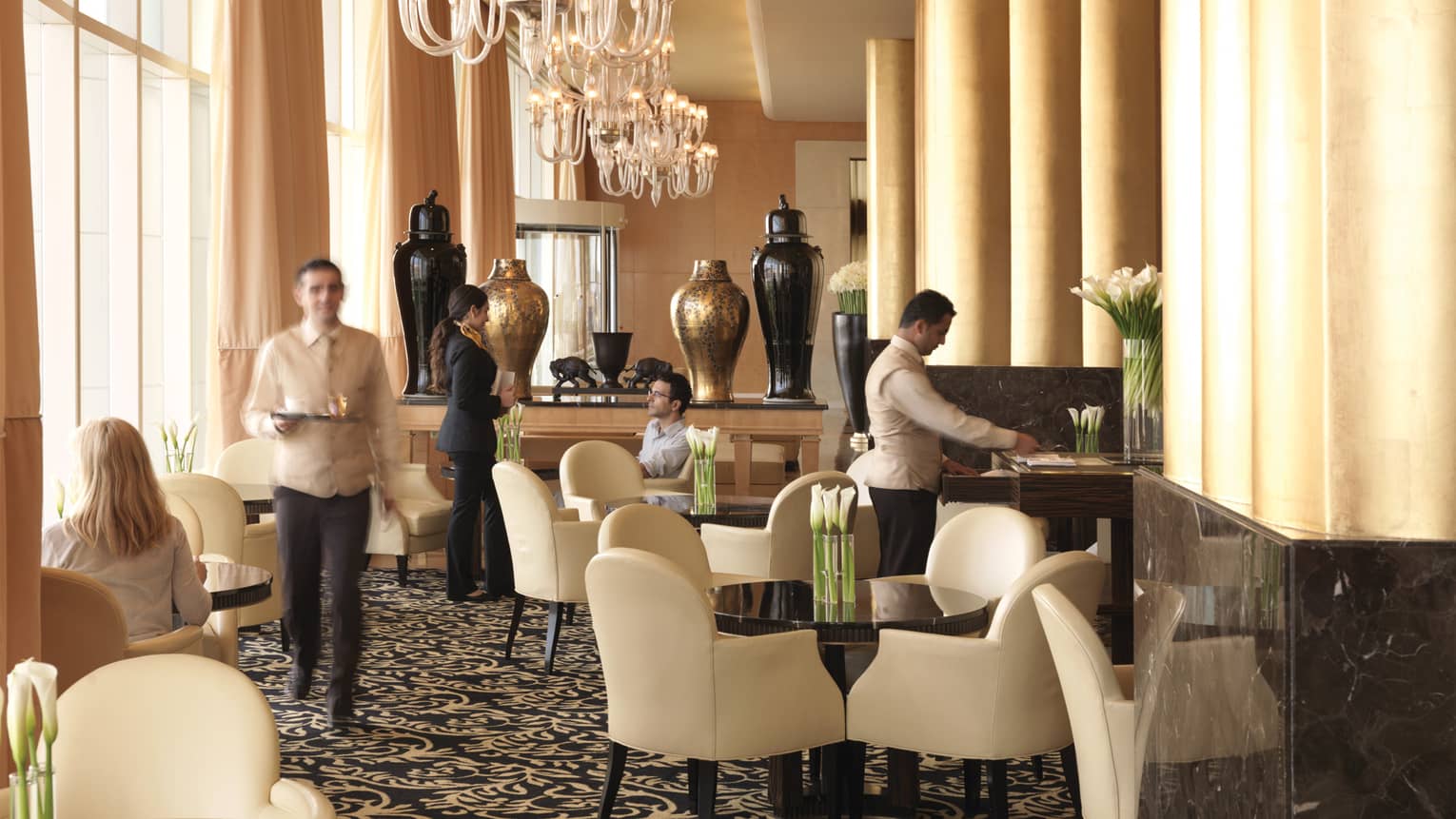 Sunny lobby lounge, hotel staff talk to guests at tables with white leather dining chairs, big gold and black vases behind