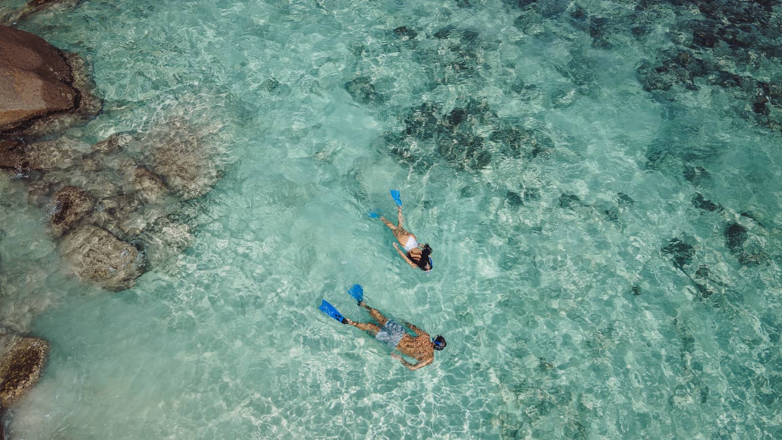 Aerial view of two snorkellers wearing blue flippers in an expanse of shallow, turquoise waters rippling over the reef.
