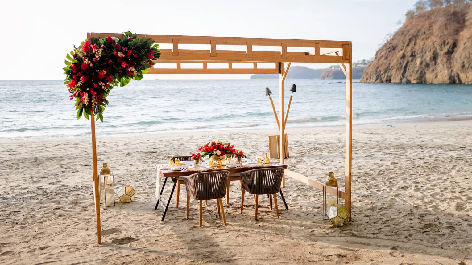 Under a pergola decorated with a vibrant bouquet of flowers, a table is set for four on the beach in the warm glow of sunset.