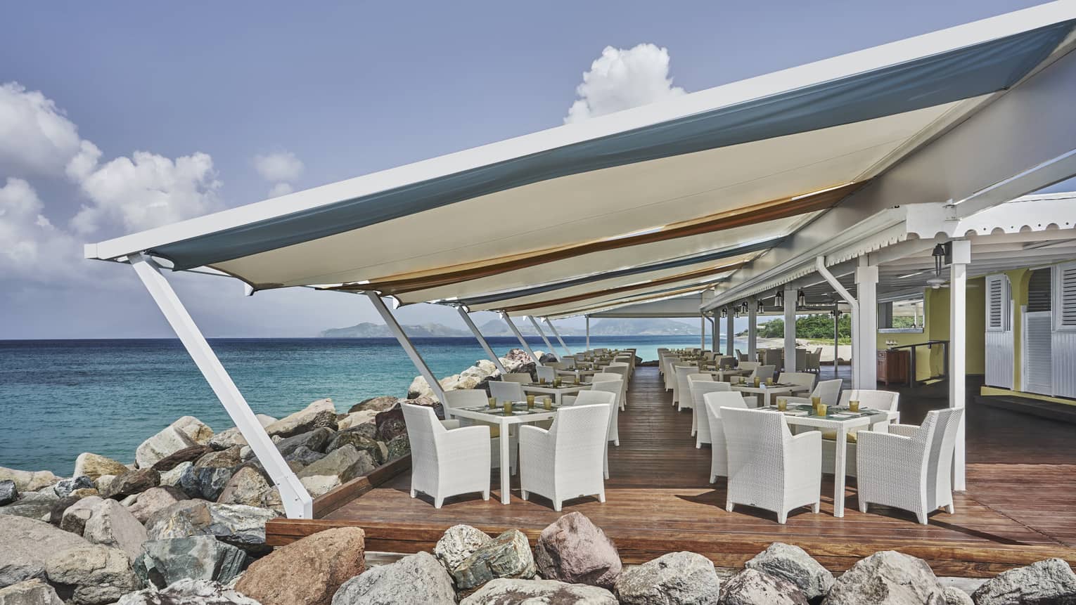 Outdoor restaurant covered by large awning, set next to large rocks and the sea