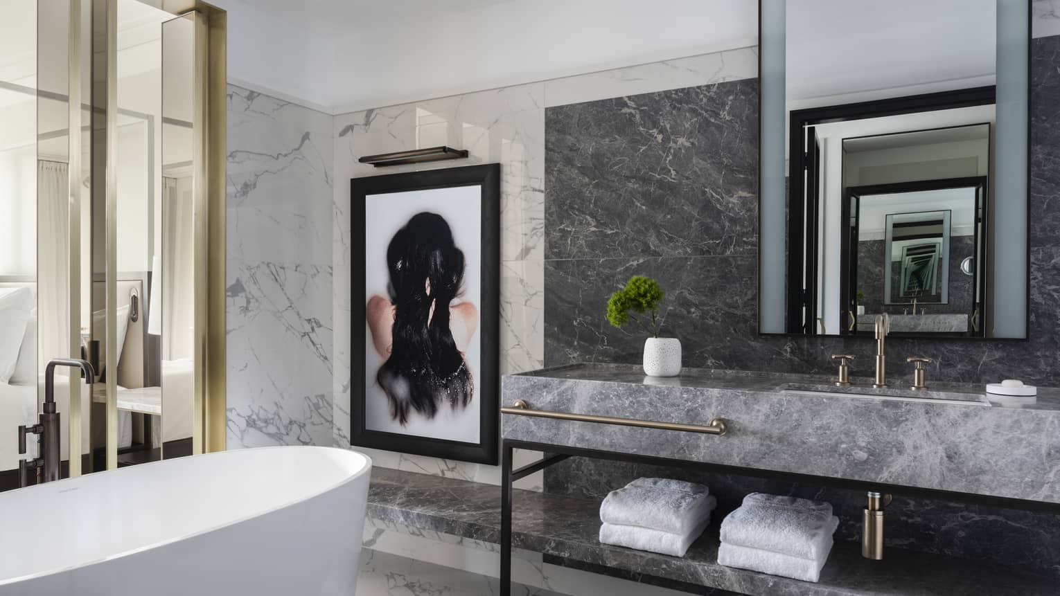 A free-standing tub, grey marble vanity and a framed work of art are set beside glass doors that open to the bedroom