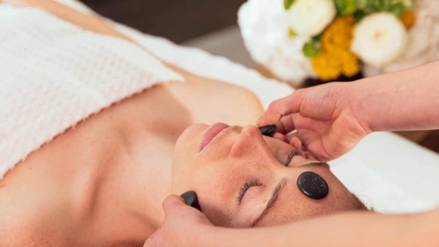 Spa staff places black stones on woman's face as she lays on treatment table
