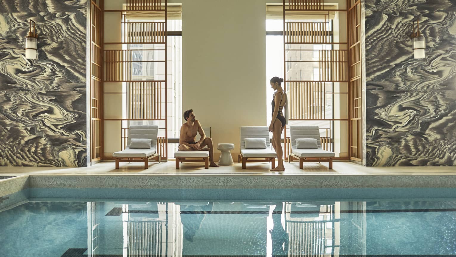 Man and woman in swimsuits by lounge chairs, window on indoor swimming pool deck
