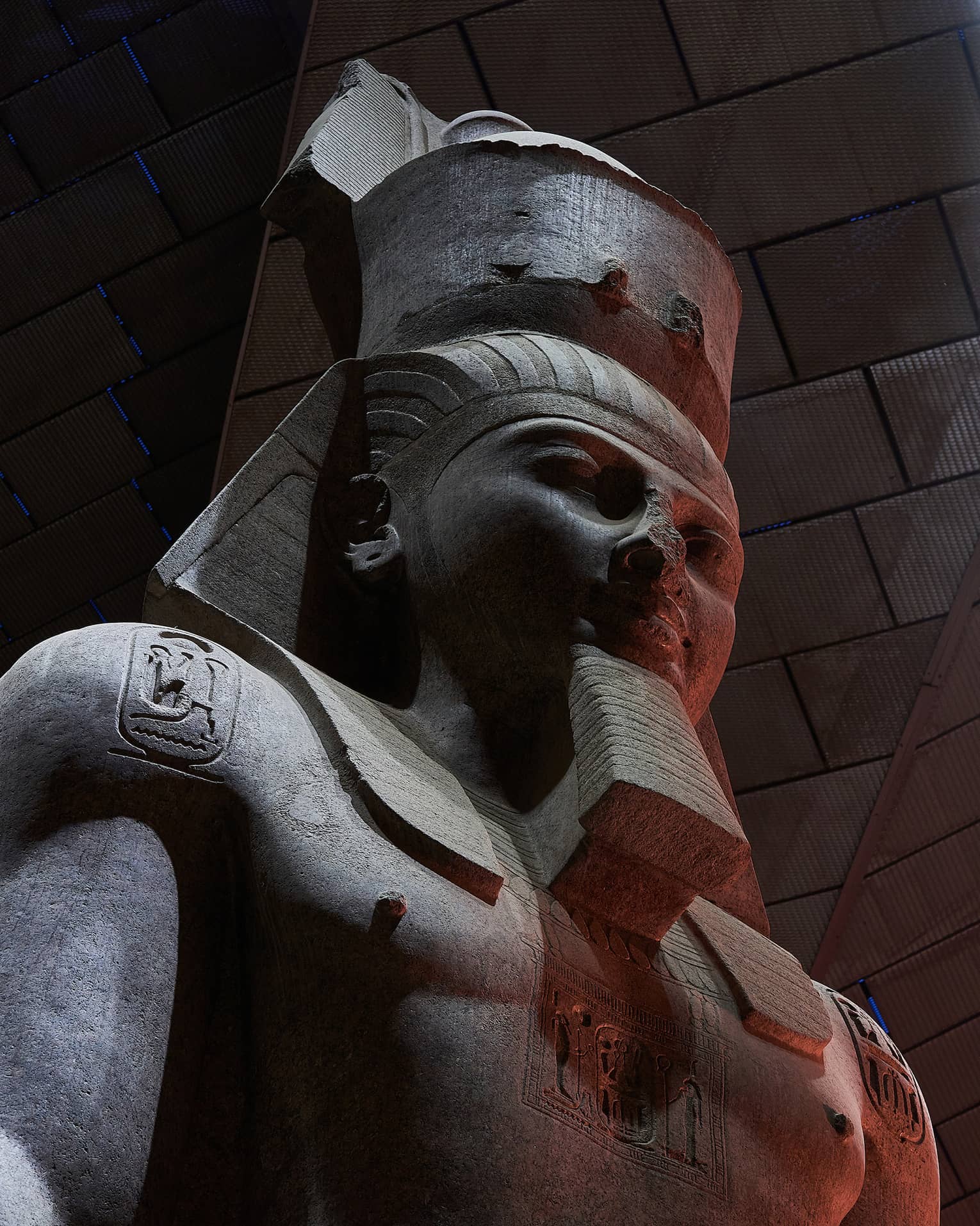 Close-up on a dark stone statue of a bearded pharoah with three heiroglyphic carvings on his chest and shoulders.