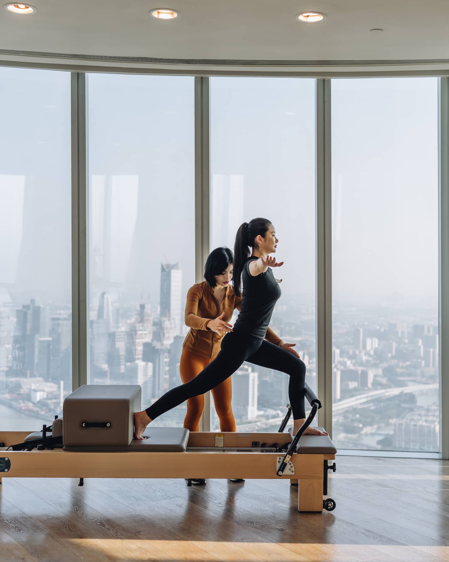 An instructor gently adjusts the positon of a guest in a lunge on a reformer machine, high up overlooking a hazy cityscape.