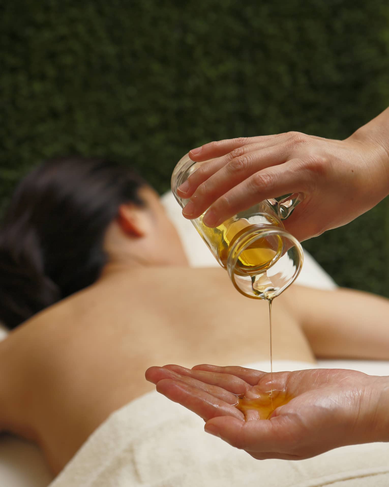 Spa staff pouring massage oil from glass jar in hand over woman lying on table