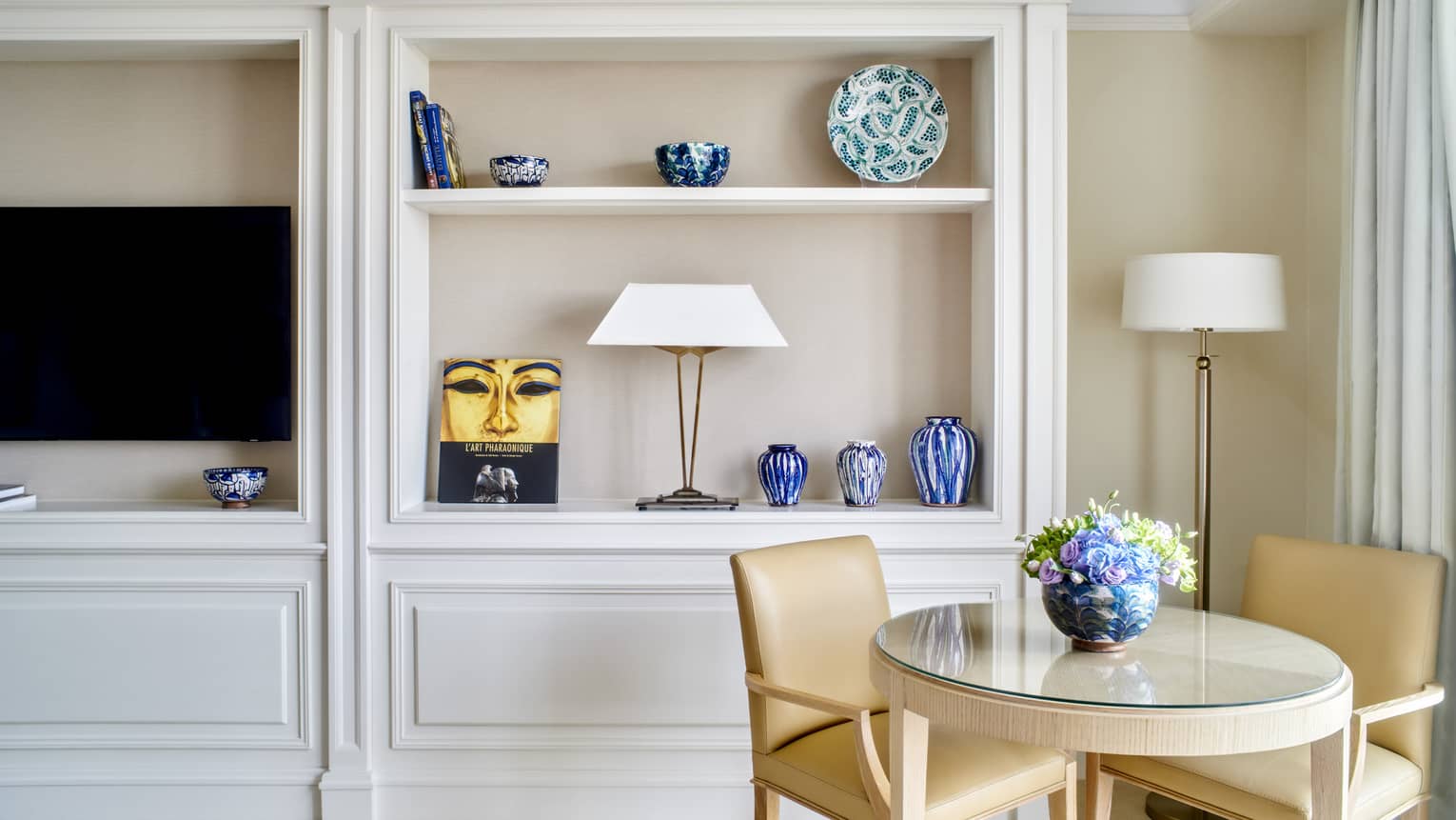 Small round table and two tan leather chairs set next to built-in wall shelves and TV