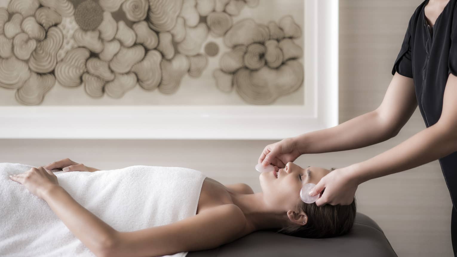 Woman lies on massage table wearing white towel as spa attendant massages her face with stones