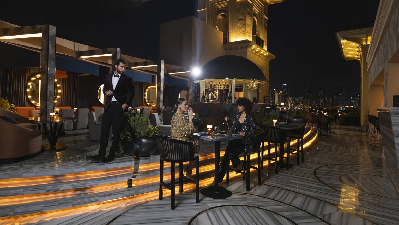 Rooftop lounge at night with two women sitting at a cocktail table and a man in a tuxedo walking behind them