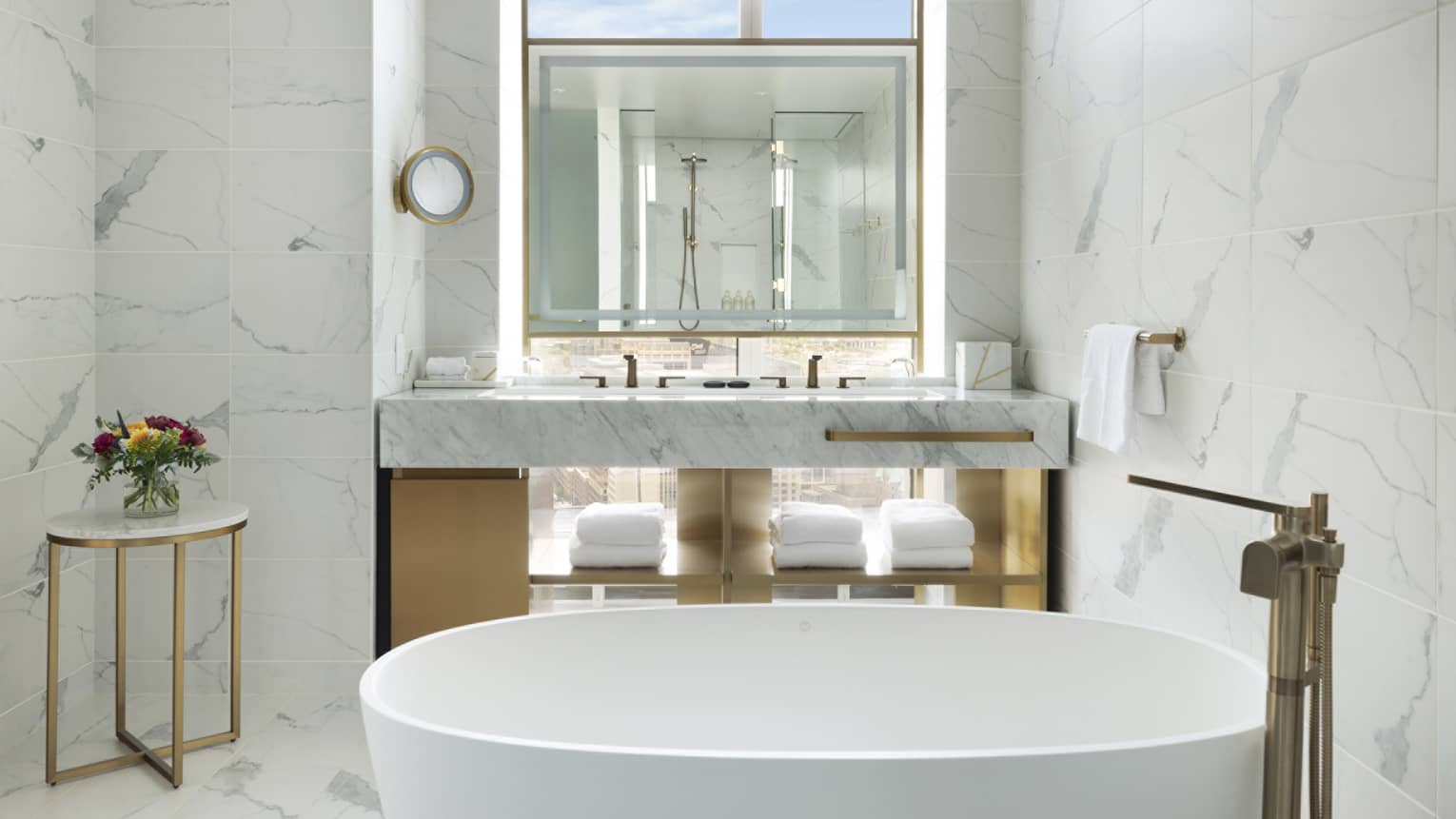 A bathroom with a deep white tub, small table, sink area with towels on shelves and a mirror.
