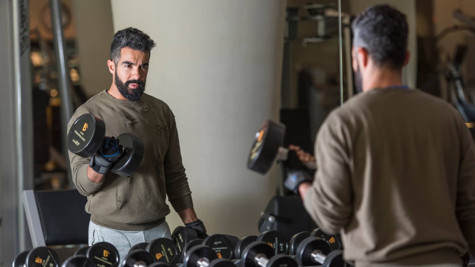 Man with beard wearing casual sweatshirt and sweatpants stands looks in gym mirror, lifts large hand weights