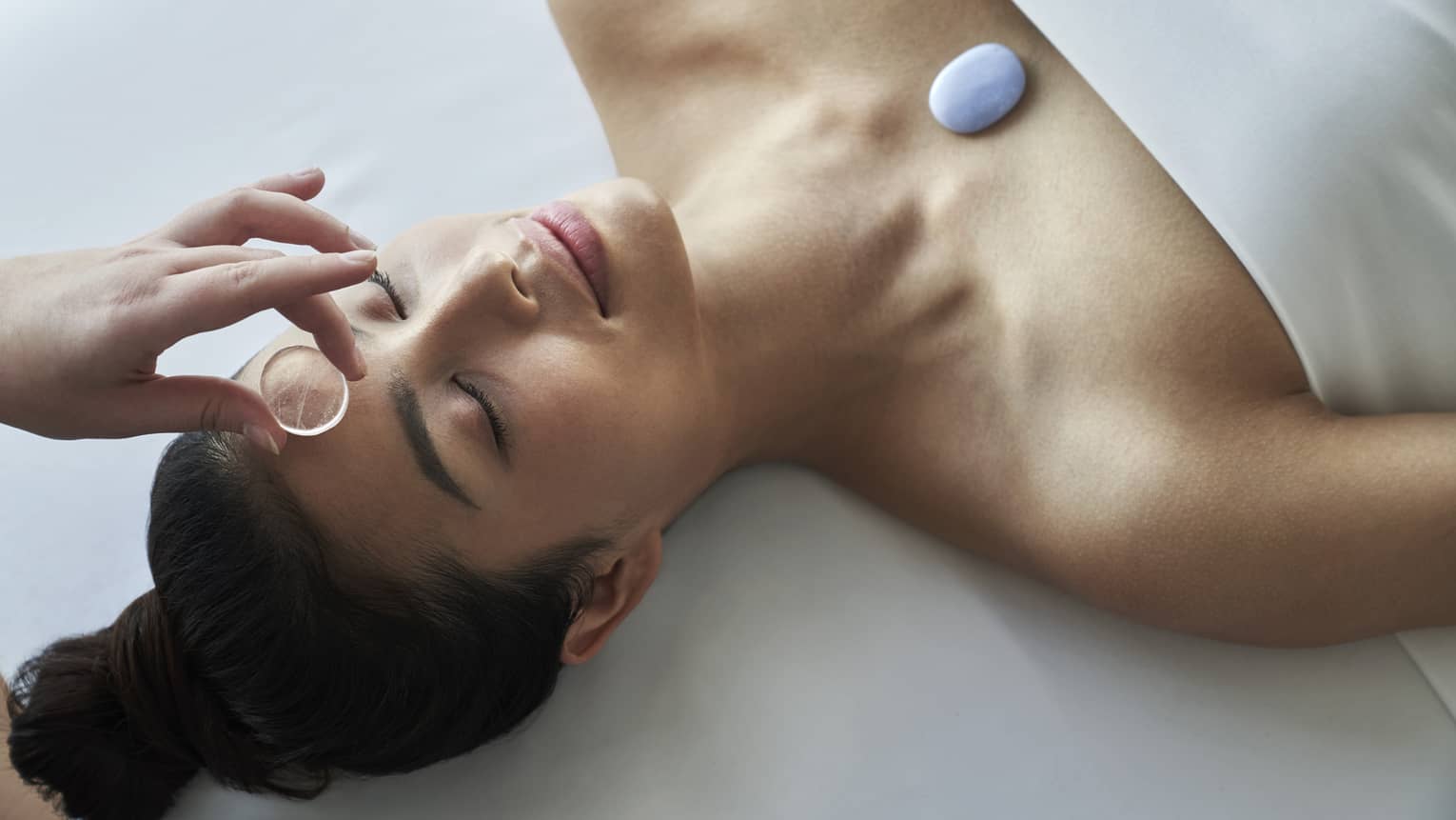 Spa staff places clear stone on woman as she lays on spa table, stone on chest