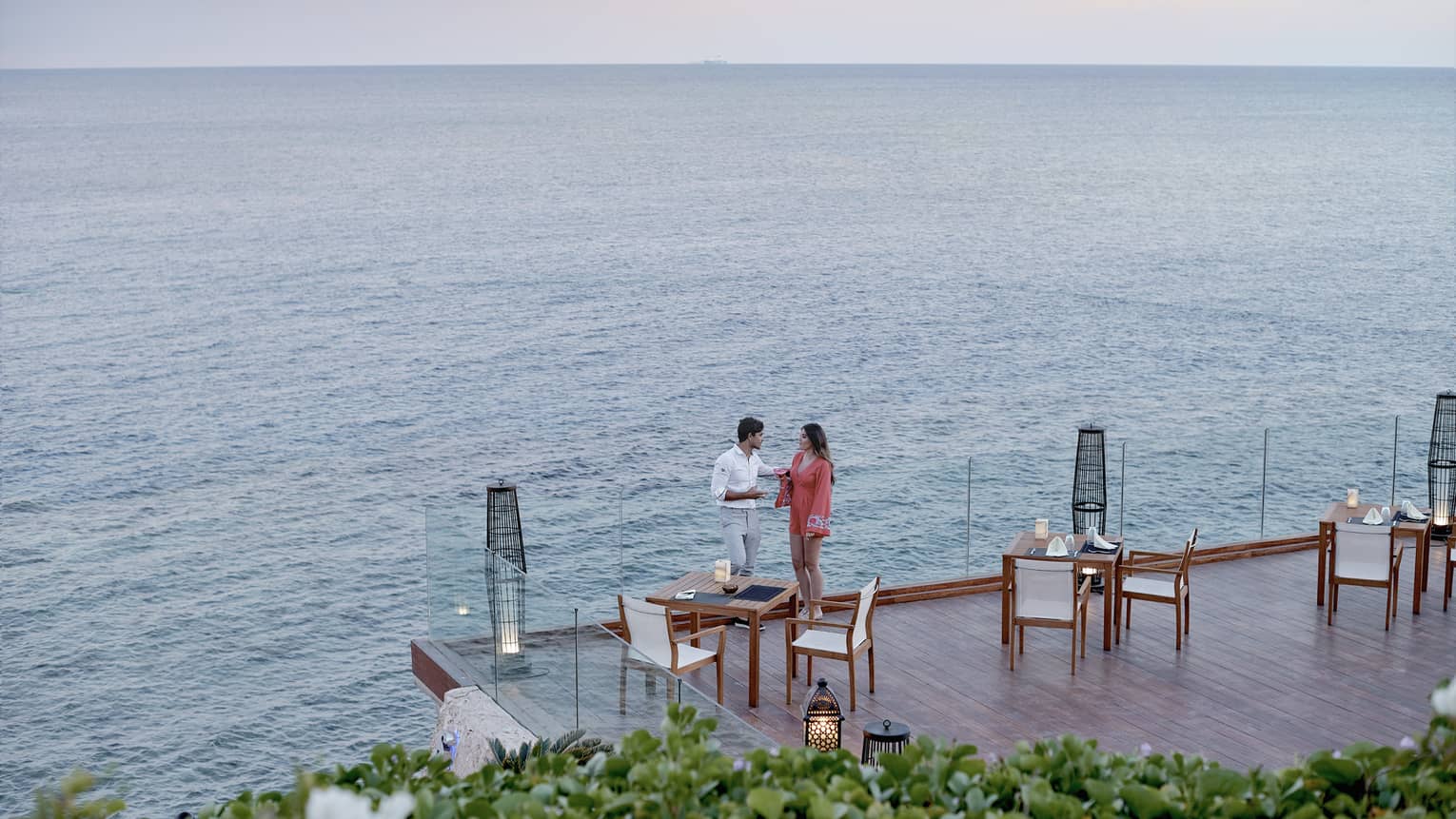 A man and woman talking on an outdoor terrace lined by glass railings, next to sea