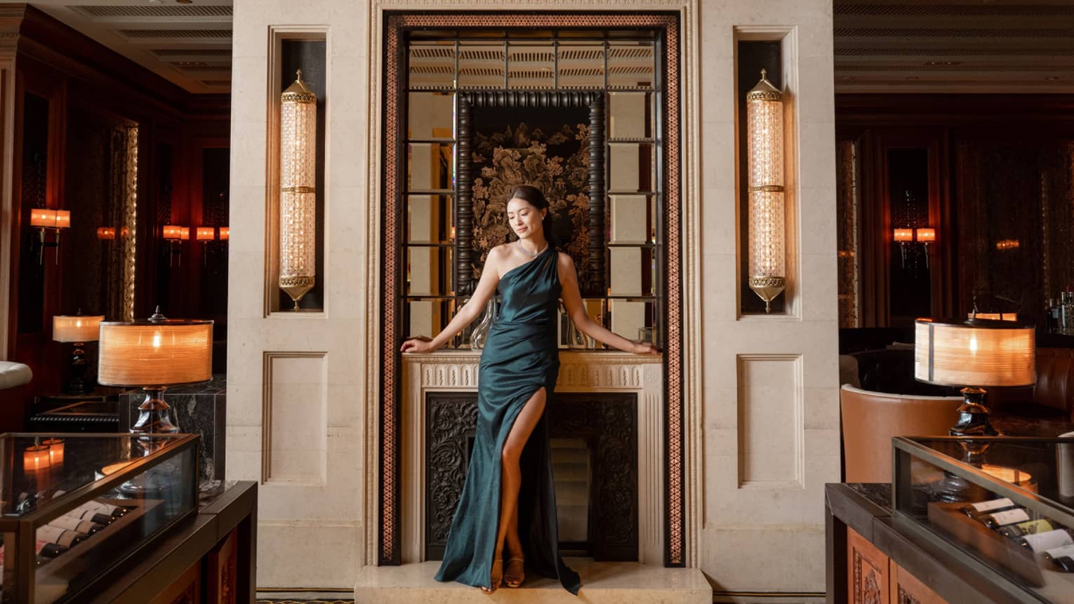 A formally dressed guest posing in front of an elegant fireplace with bottles of wine in tabletop glass cases on either side.