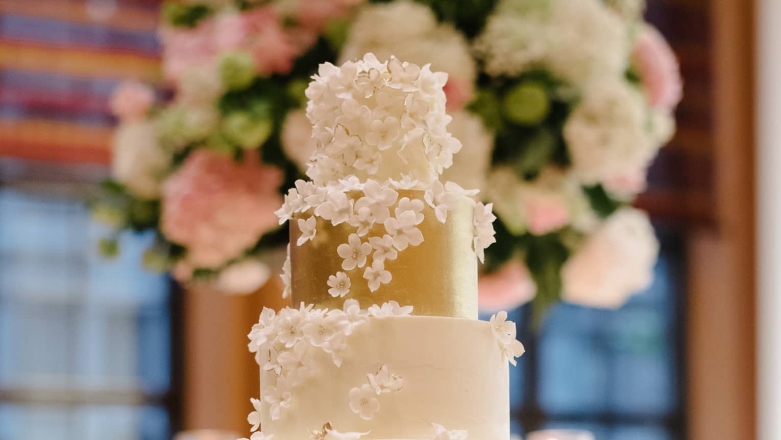 Five tiered white wedding cake with gold layer, white icing flowers on table with tealight candles