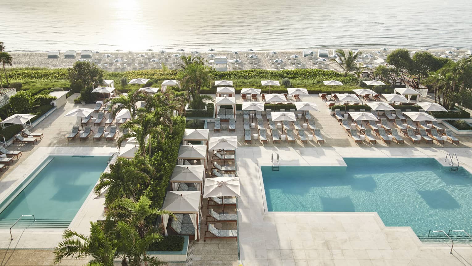 A pool area near a beach with many lounge chairs and umbrellas, surrounded by palm trees.