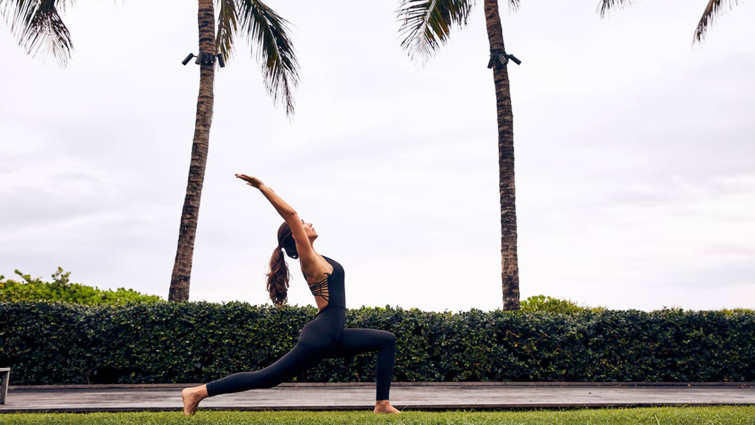 A woman outside on grass next to palm trees doing yoga.