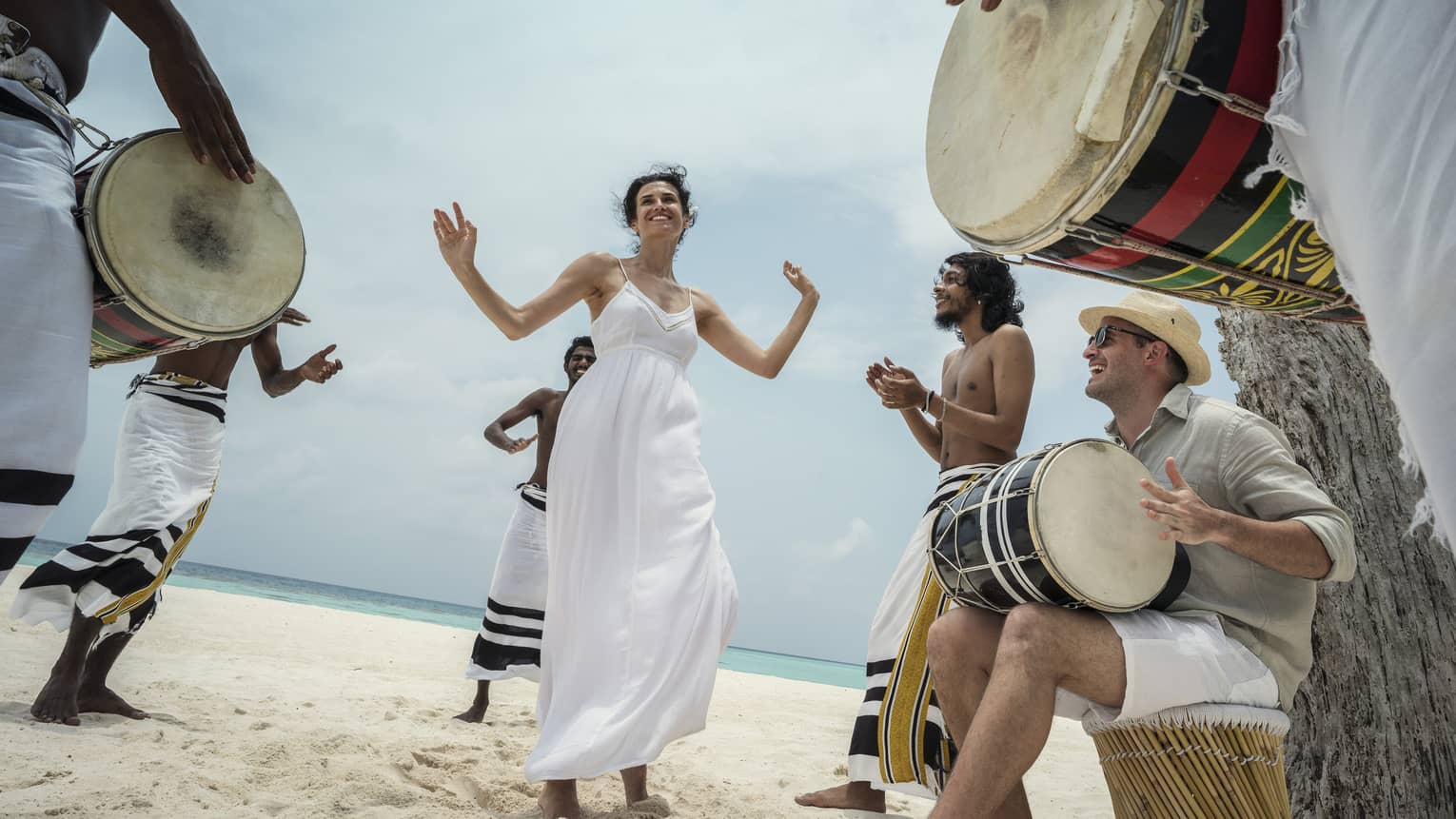 Woman wearing white dress dances in middle of drummers on beach