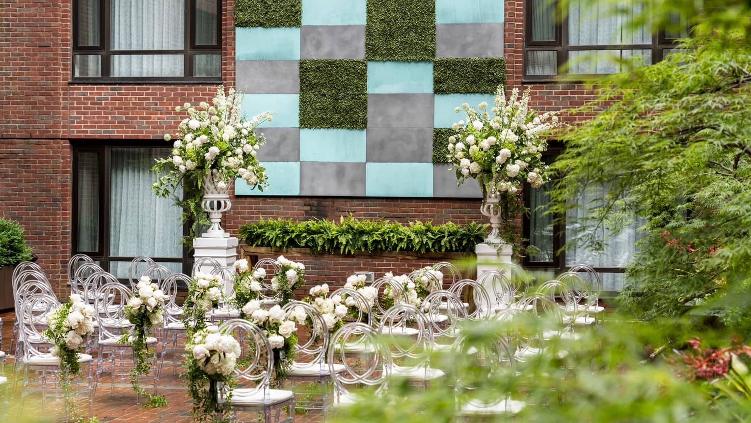 An outdoor area with rows of white seats and flowers arranged around them, a brick walled building can be seen in front of the chairs.