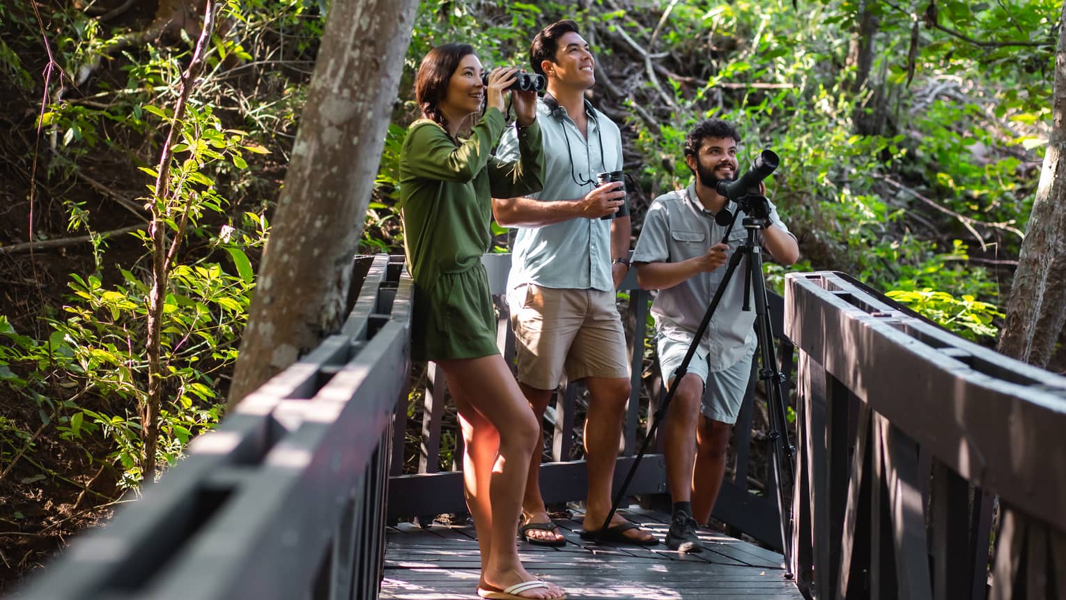 A woman holding binoculars, a man holding a recorder and a guide holding a camera on a tripod are gathered on a wooden forest walkway for bird watching