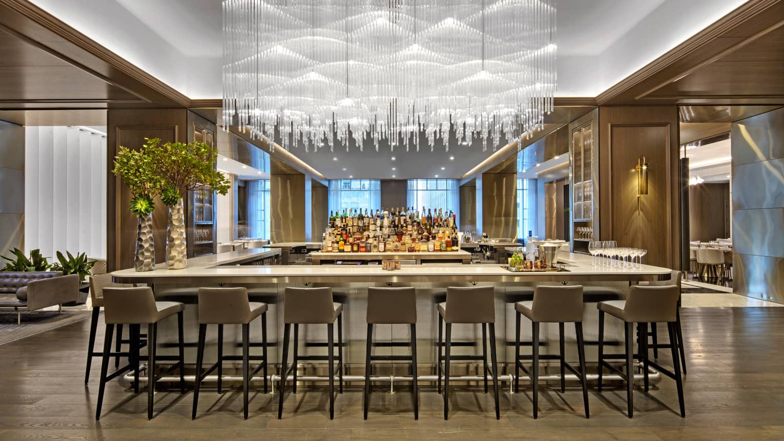 A large bar area with hanging glass chandelier and many grey stools.