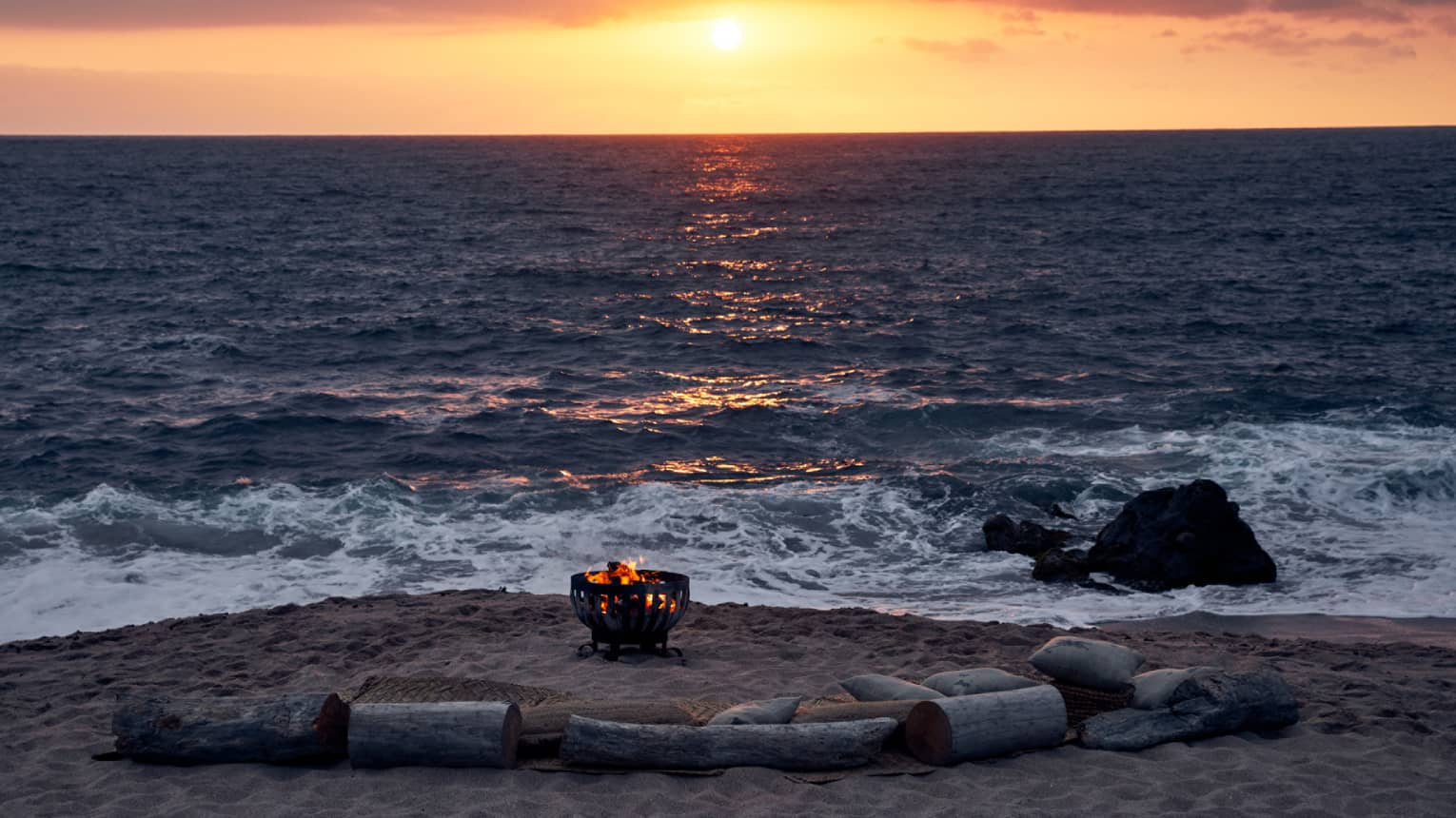 Fire burns in an iron pit on the beach, semi-encircled by driftwood log seats facing the sun setting into the ocean horizon.