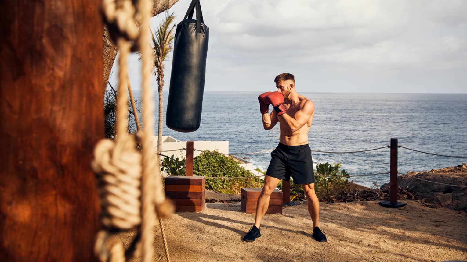 A man in boxing gloves near a punching bag and the ocean.