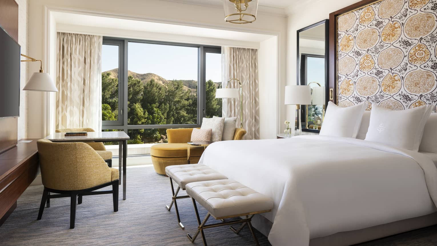 Guest room with white King bed, mustard-coloured chaise lounge & chair, and a view of trees 