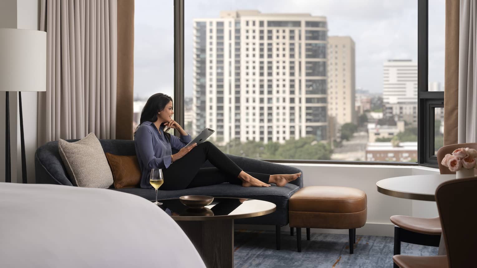 A woman sitting on a lounge chair by a large window reading.