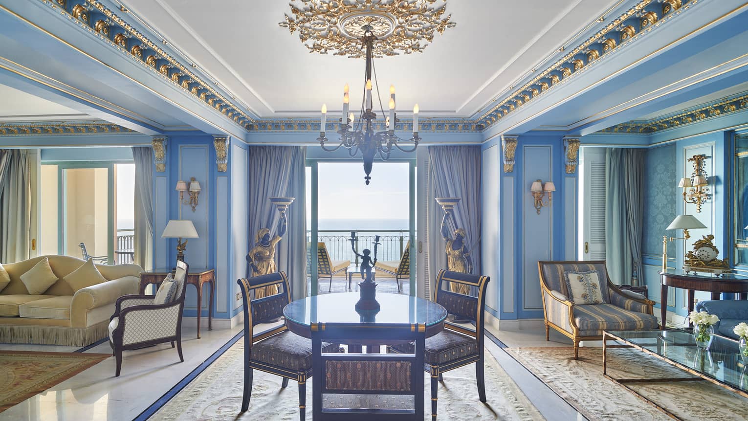Presidential Suite dining/living area with round dining table, chaise longue, cream couch, blue wall accents, doorway to veranda with sea views