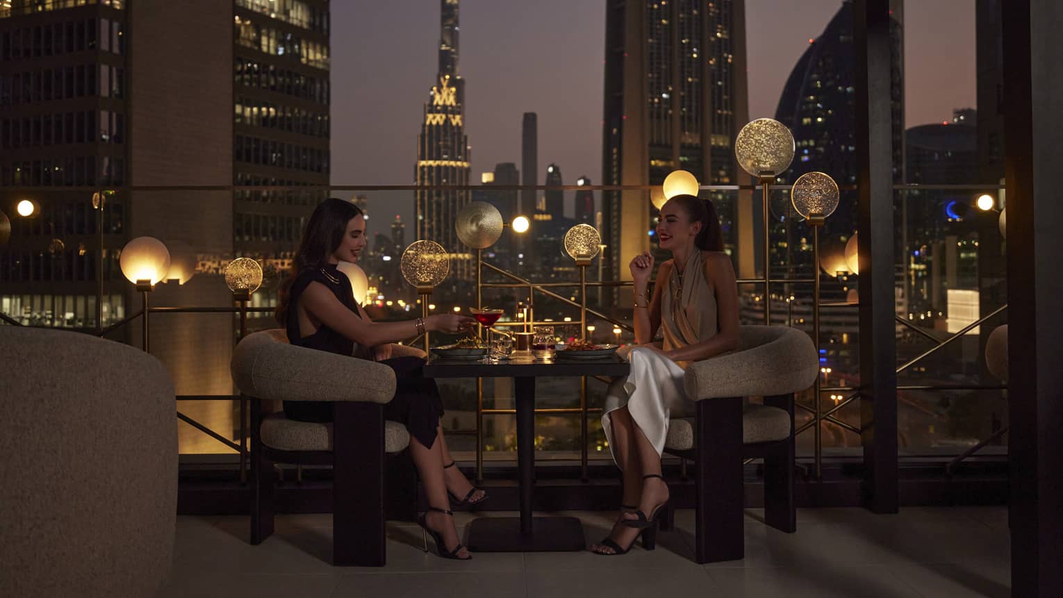 Two women sit at a cocktail table on an outdoor rooftop patio overlooking the Dubai skyline at night