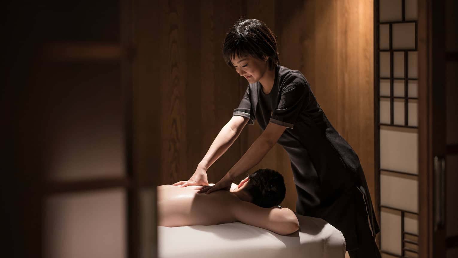 Man lies face-down on massage table as masseuse in black uniform rubs his shoulders