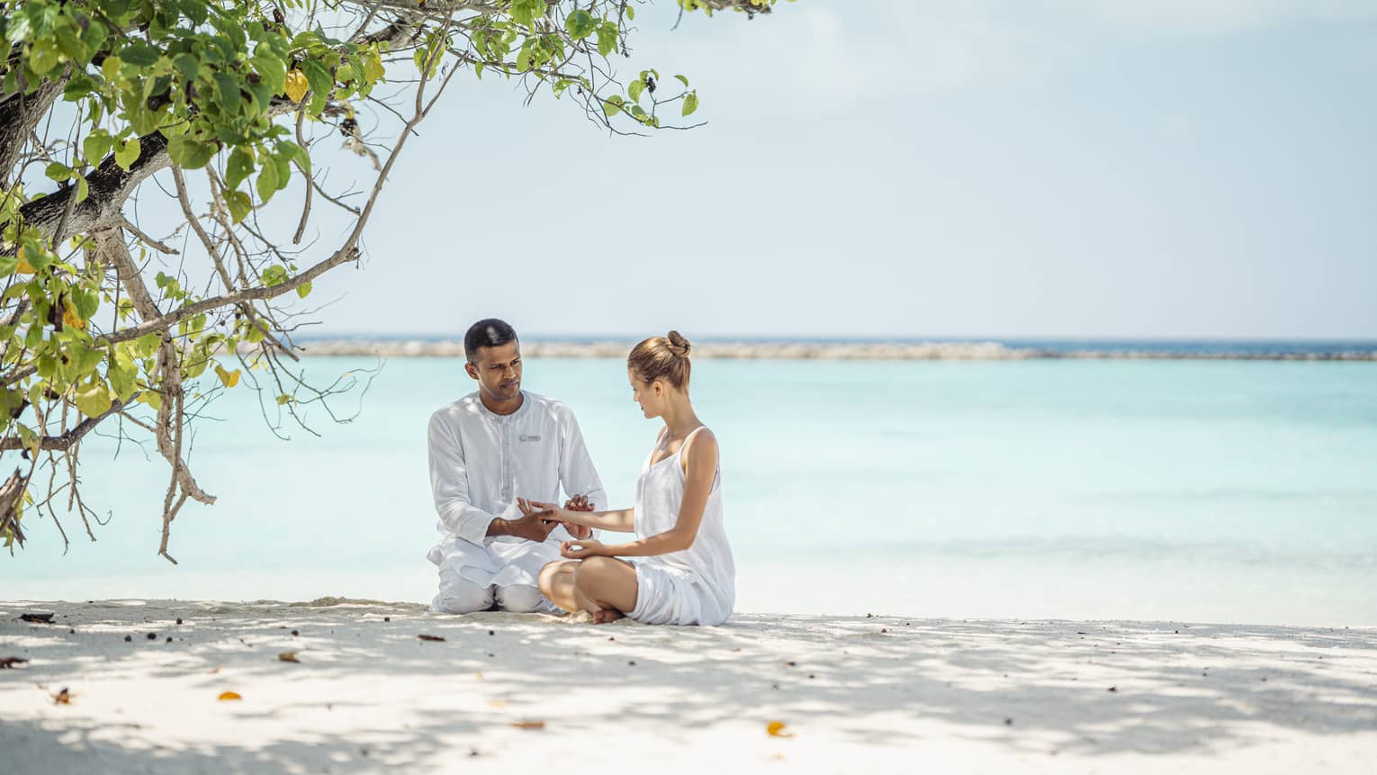 Dr. Arun Tomson teaches holistic practices to female guest on the shore, crystal-clear blue water in the background