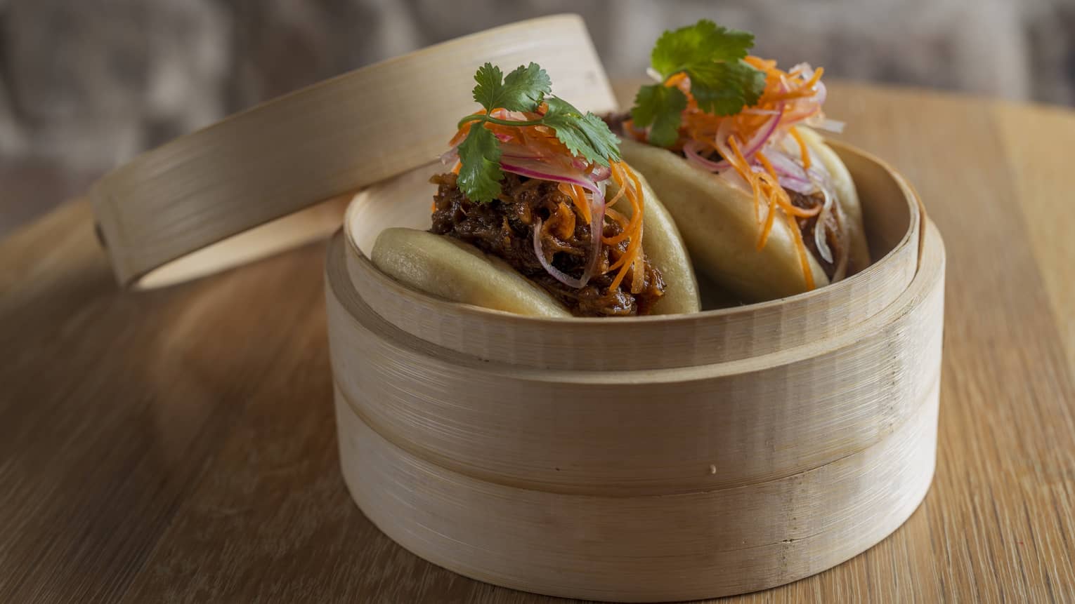 Two Asian baos with slow-braised pork ribs, wasabi mayo and pickled radish salad served in a bamboo steamer basket