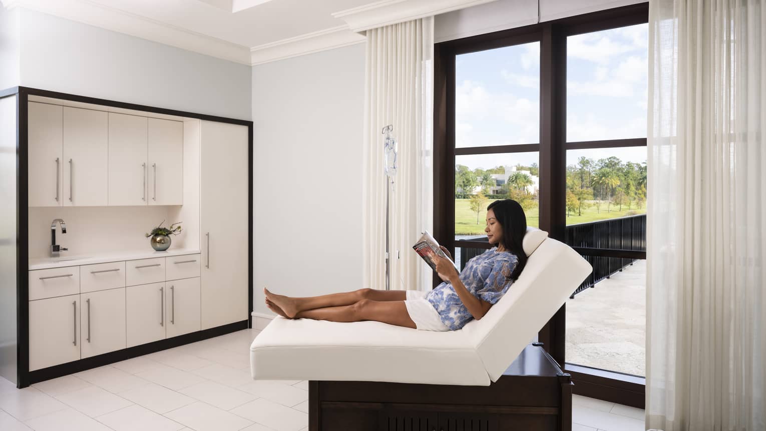 A woman reading a magazine on a folded mattress in a spa clinic with large window.