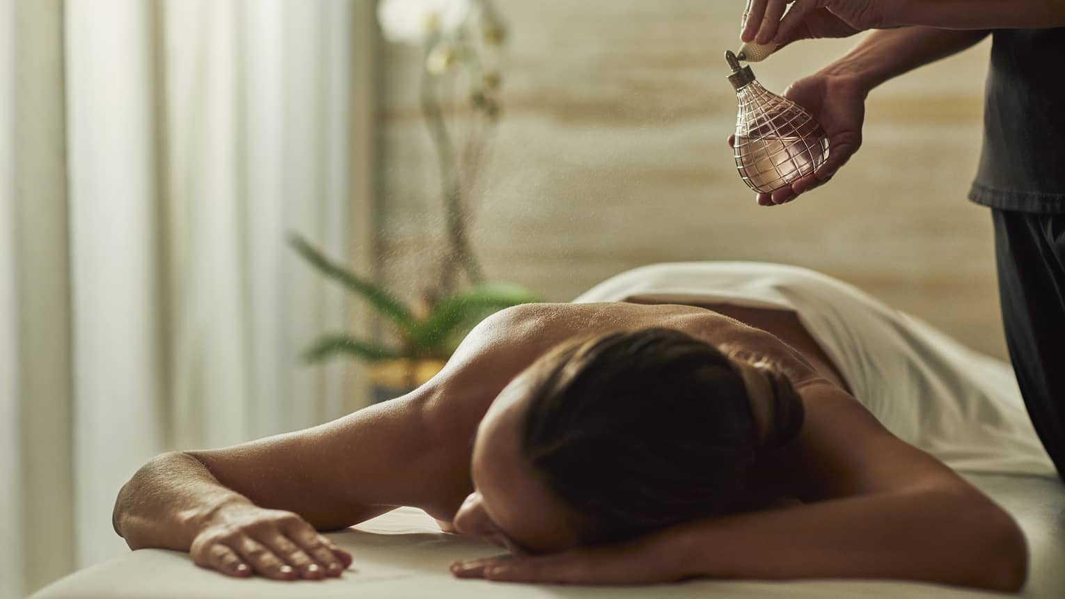 Spa staff sprays perfume bottle over woman lying on massage table
