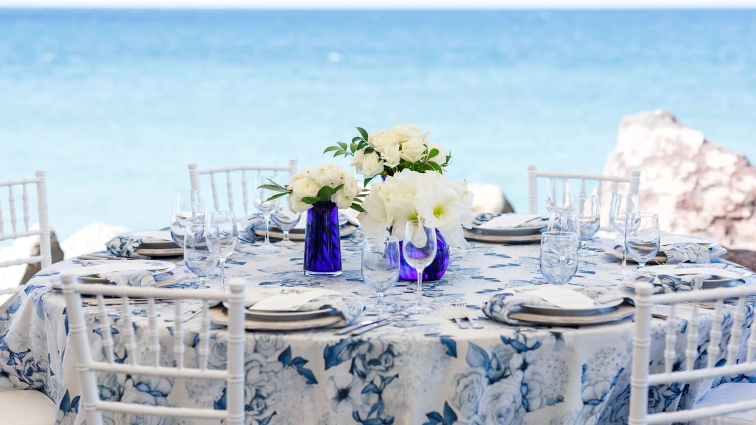 Blue and white round table formally set, five white chairs, white floral centrepieces, on beach