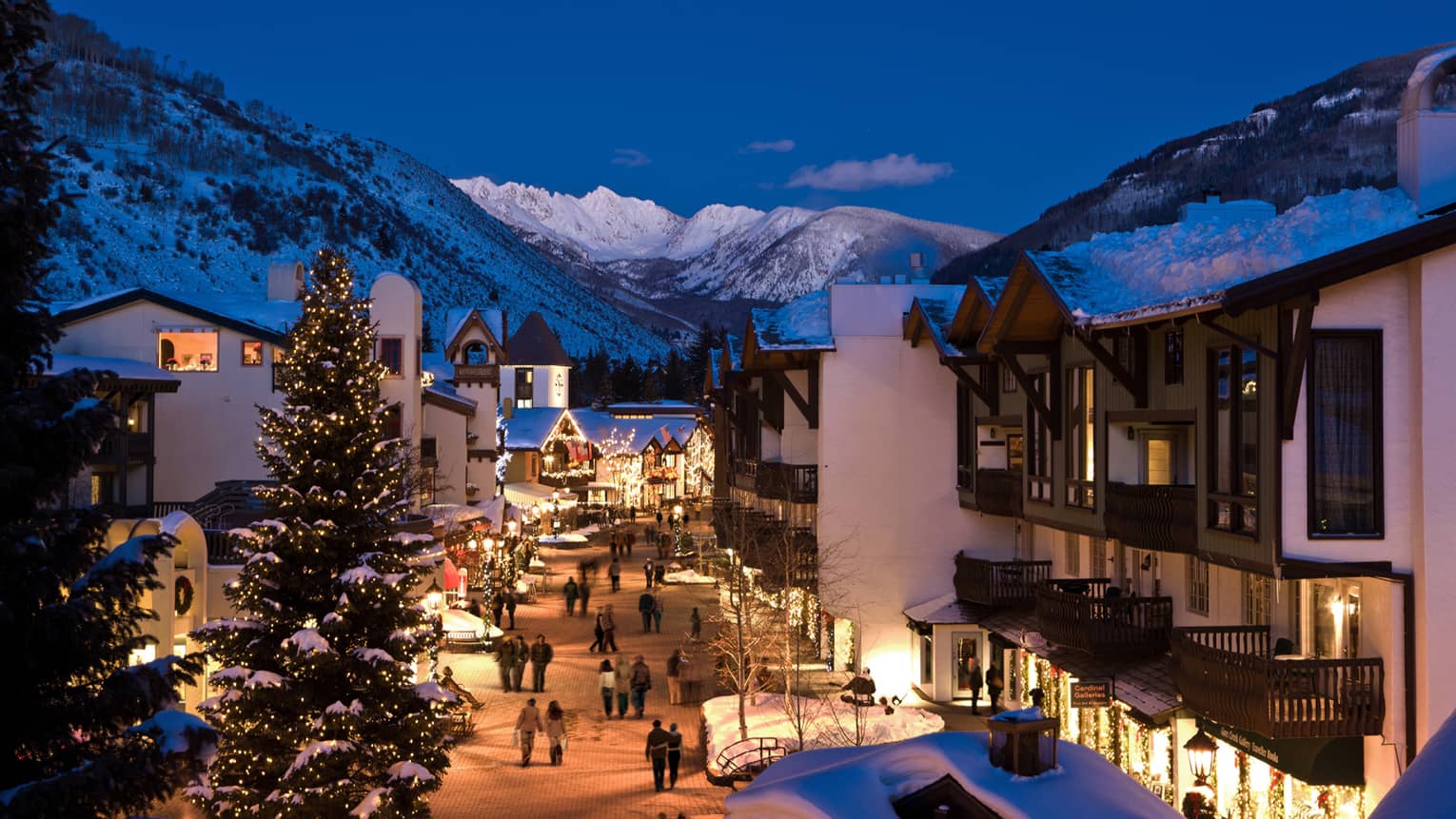 A snow covered hotel property surrounded by mountains and trees with lights coming out of buildings and street lamps at night.