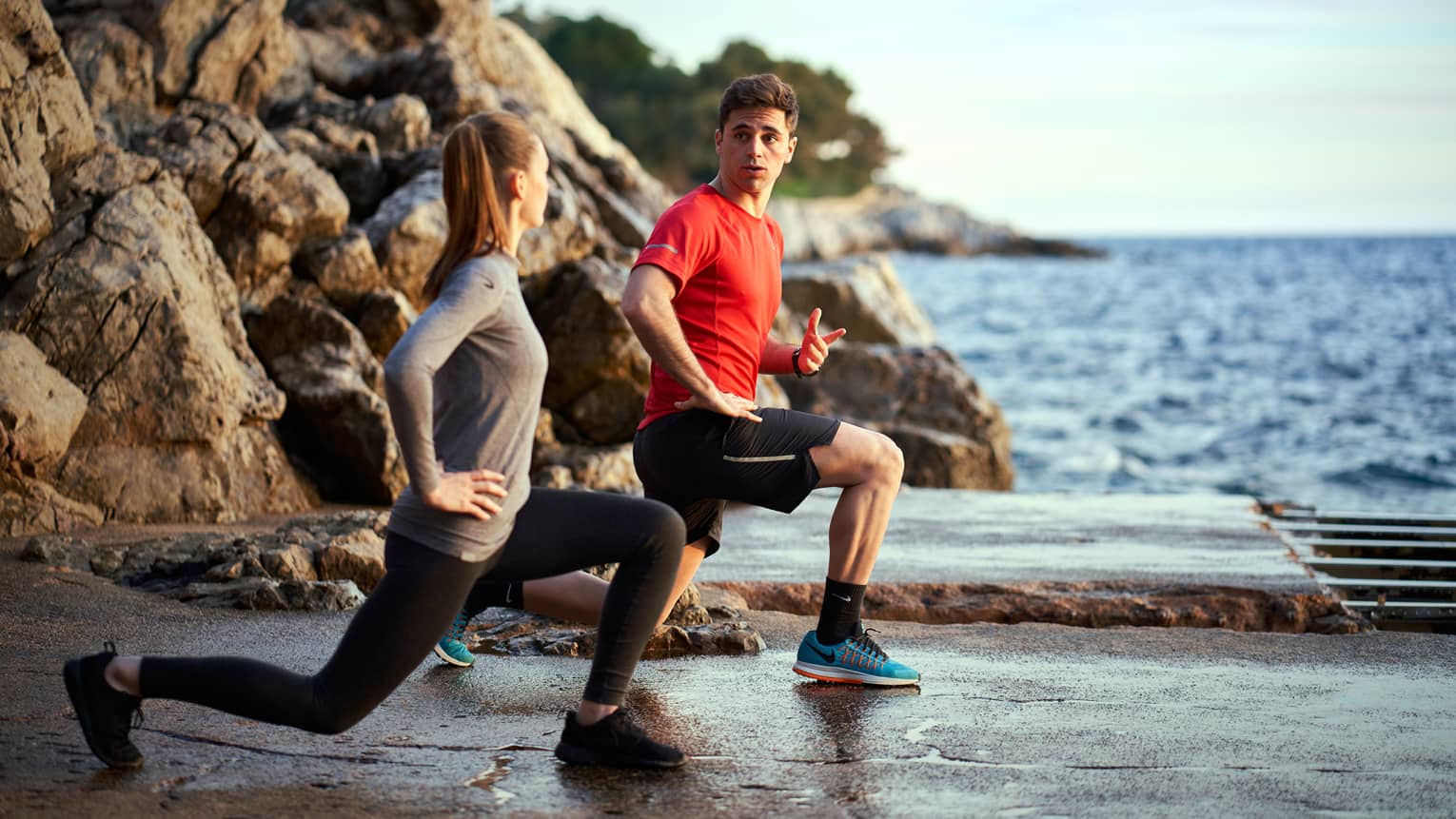 A man trains a woman while doing lunges outside next to the sea and rocks