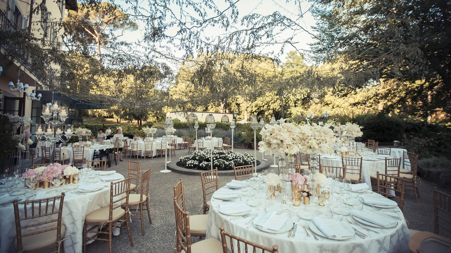 Circular tables are set with white porcelain dishes, silverware, pink and white florals in a courtyard surrounded with brush and sting lights