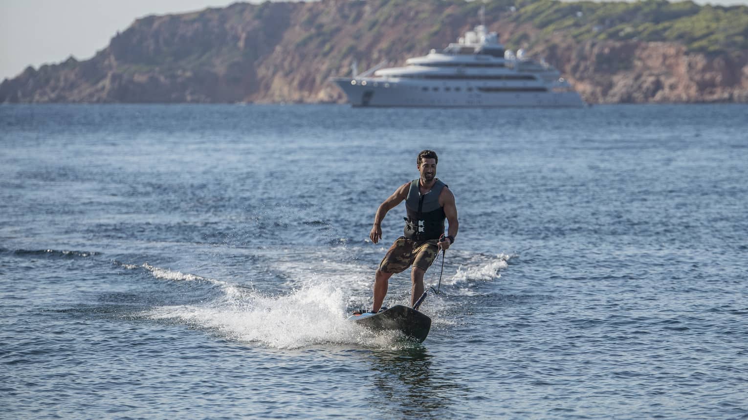 Front view of a guest jetting along the sea on a motorized surfboard, a majestic ship and forested mountains in the backdrop.