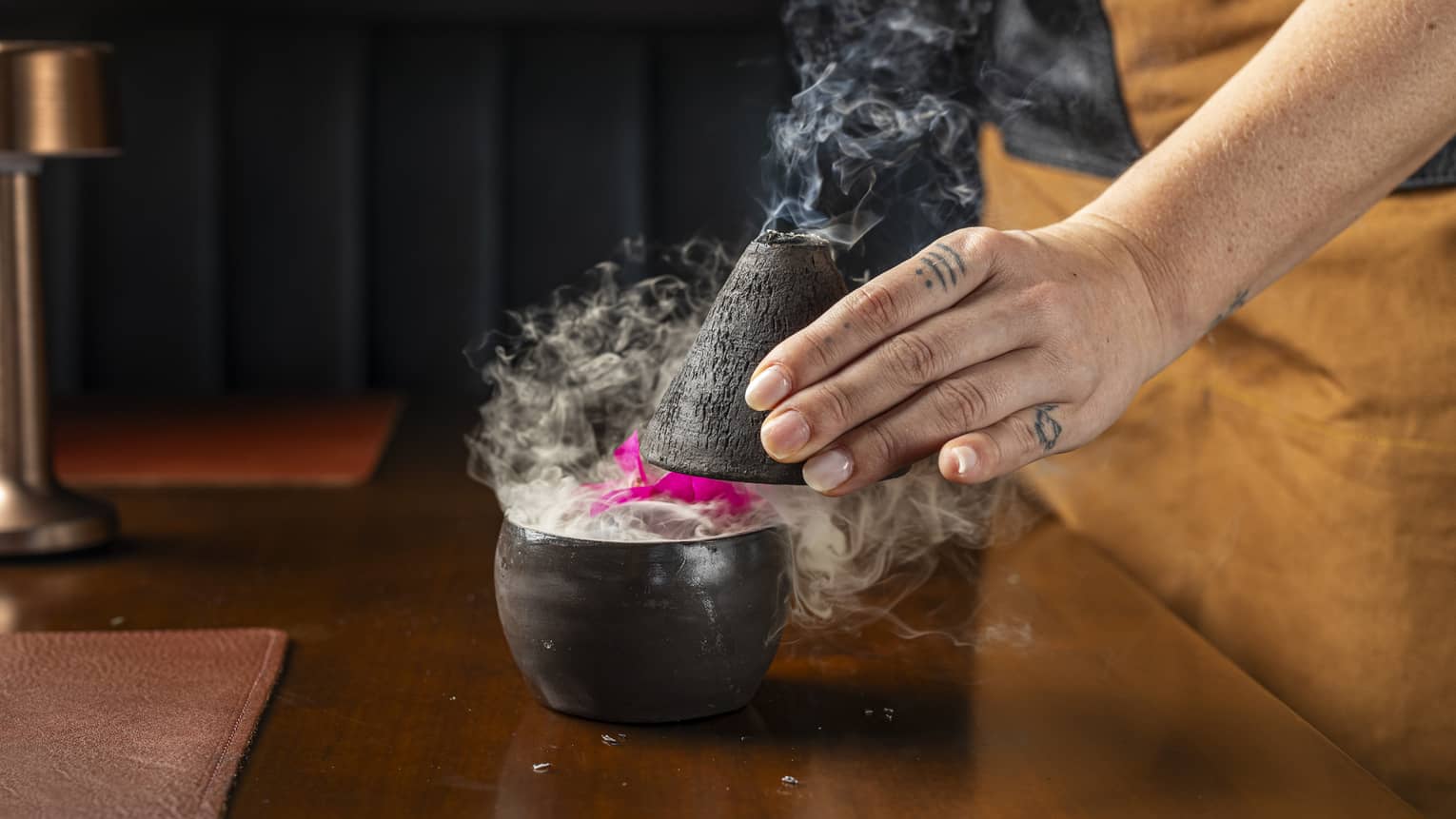 A tattooed chef's hand removes a black cone top from a small black bowl revealing a bright pink flower and plumes of smoke