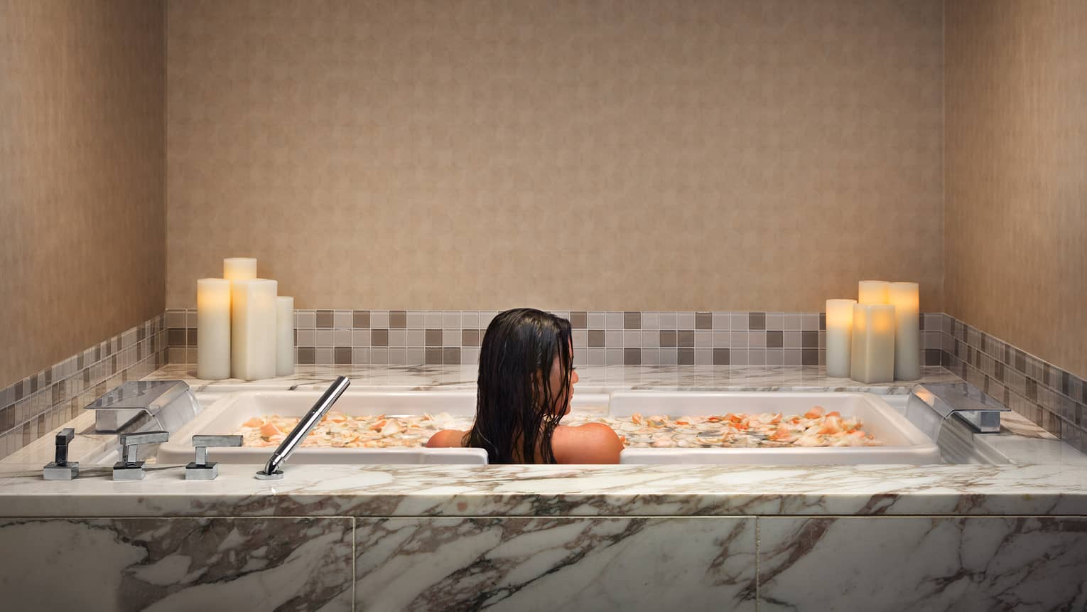 Woman soaks in marble tub filled with flower petals and lined with candles