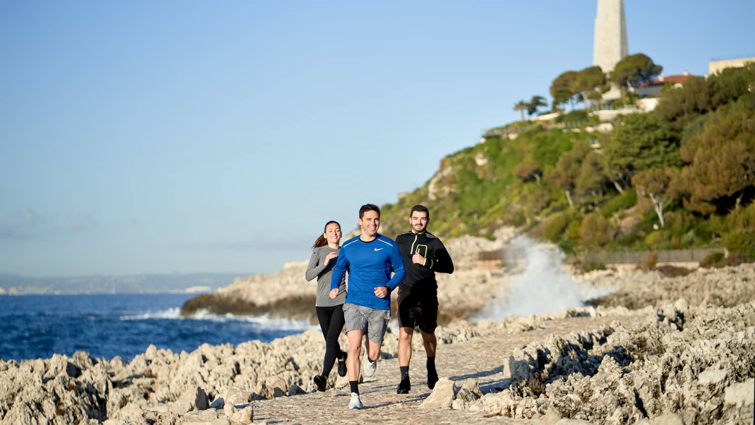 Three people jogging, smiling, on rocky trail beside the sea, lighthouse in backdrop