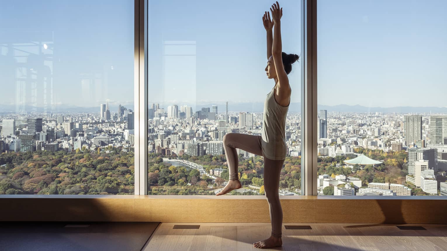 Woman stands in yoga pose in room with floor-to-ceiling windows with city views