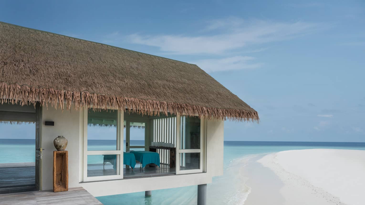 Exterior view of thatched roof spa with massage beds in open-air room over lagoon