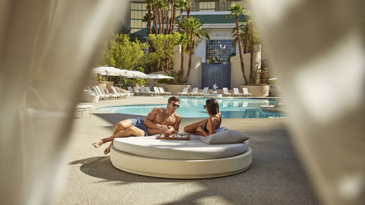 A man and woman lounging on a round daybed by a pool.