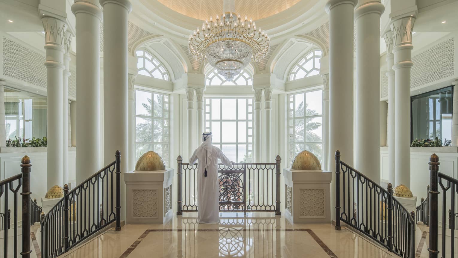 A man in a white thobe in front of enormous floor-to-ceiling windows in a marble foyer. 